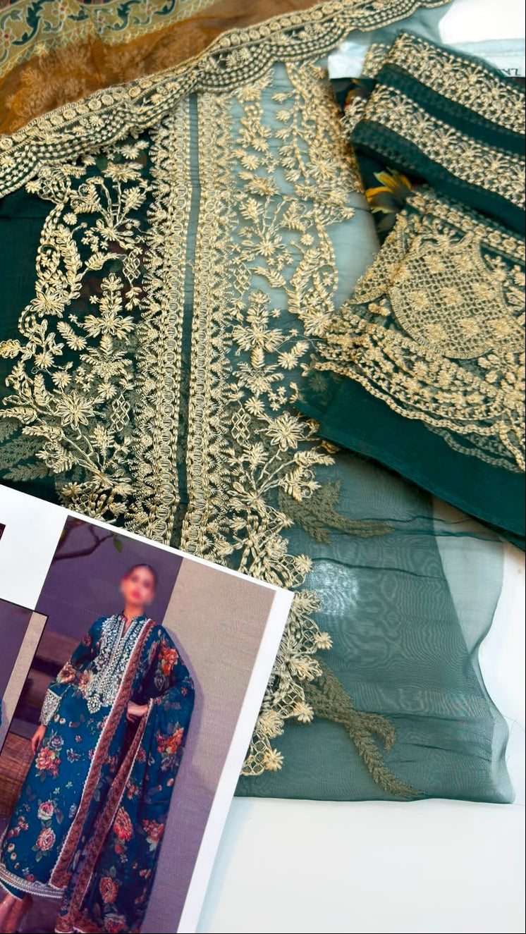 A folded embroidered ethnic outfit fabric set in deep green with gold floral embroidery and a matching sheer dupatta, displayed alongside a printed fashion magazine on a white surface