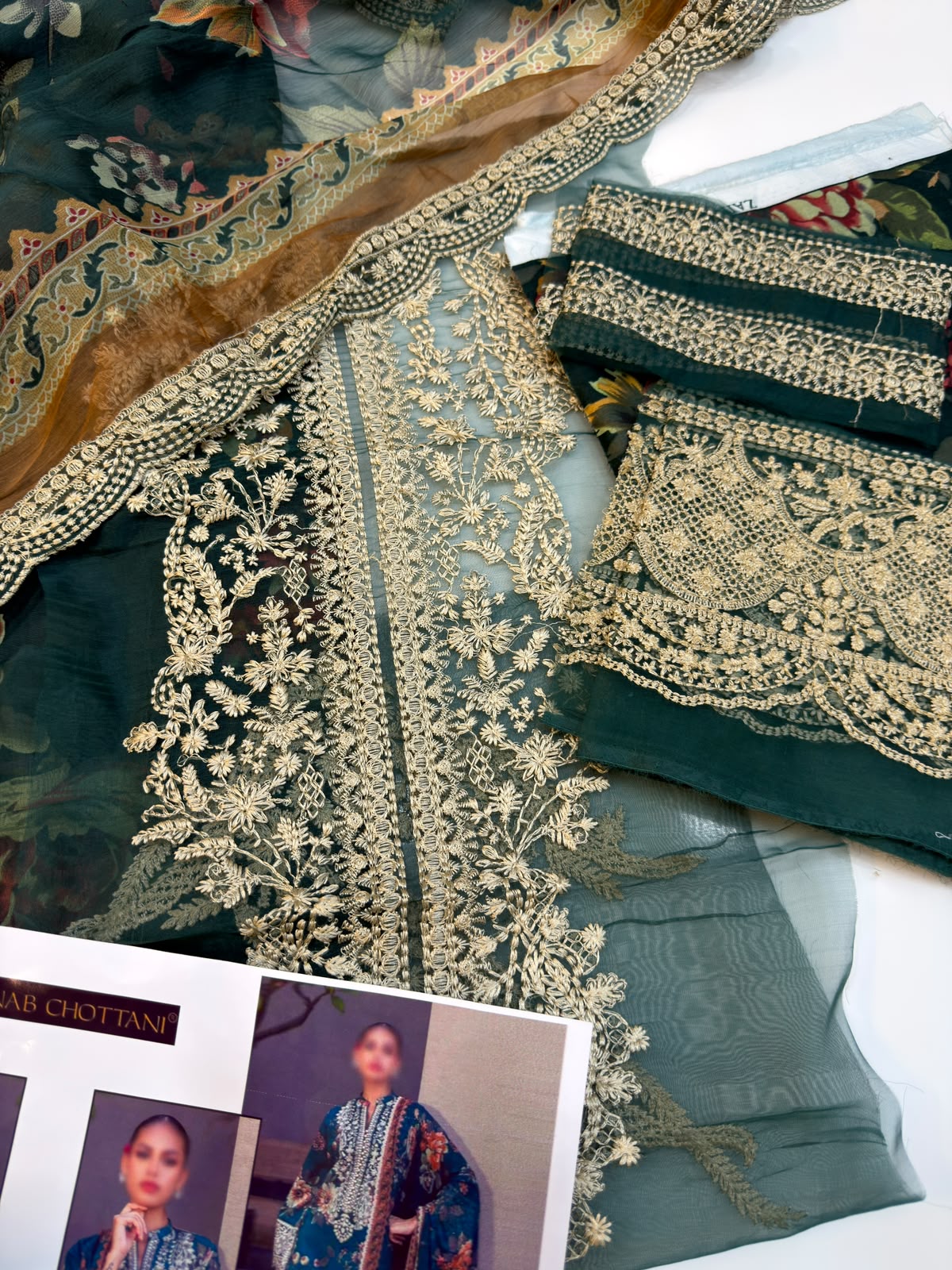 intricate gold floral embroidery, matching embroidered borders, and a coordinating dupatta. A printed magazine or catalog page with photos is partially visible at the bottom.